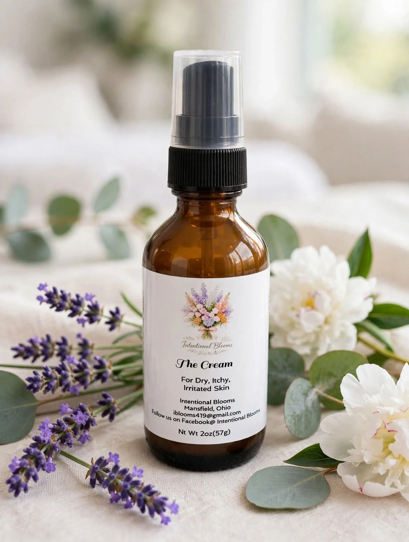 A small amber glass spray bottle with a black spray nozzle and clear cap, surrounded by sprigs of lavender, white flowers, and eucalyptus leaves on a light fabric surface.