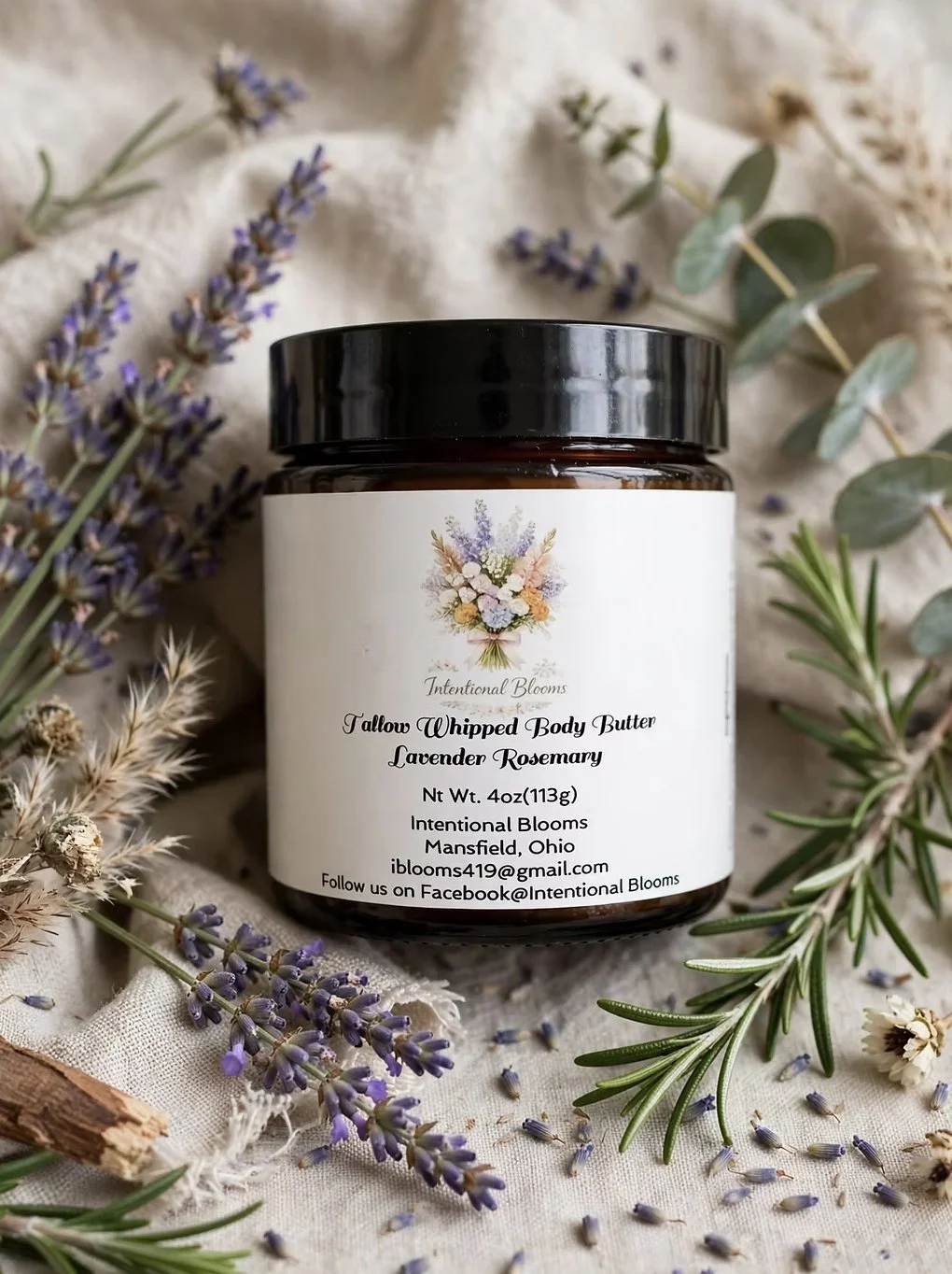 A jar of lavender rosemary scented body butter surrounded by sprigs of lavender, rosemary, and other dried herbs on a cloth background.
