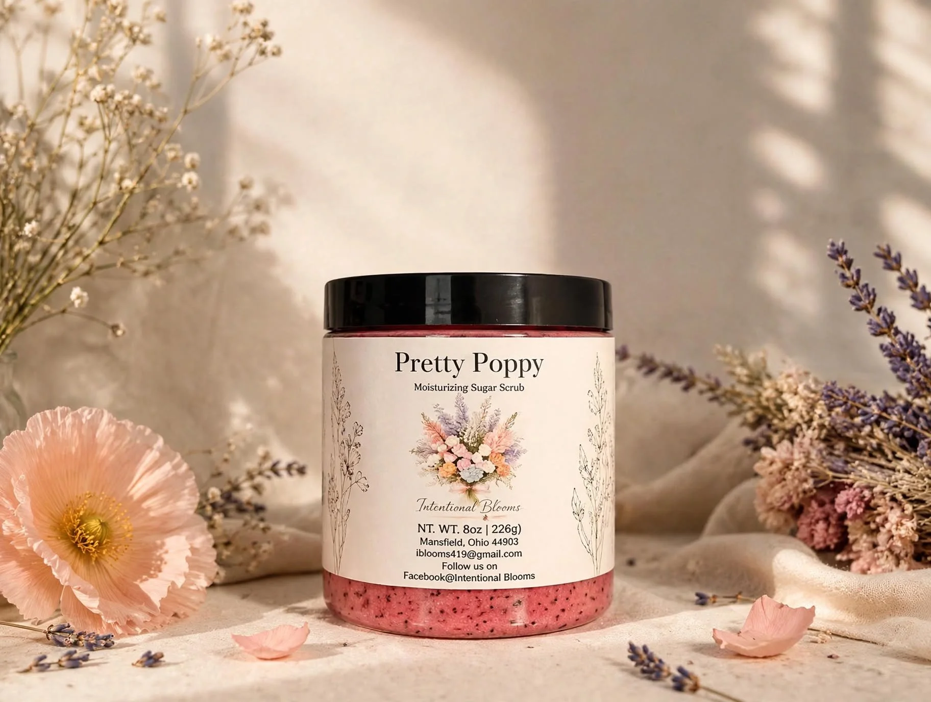 A container of Pretty Poppy Moisturizing Sugar Scrub labeled 'Intentional Blooms' surrounded by pink and purple flowers on a soft fabric background.