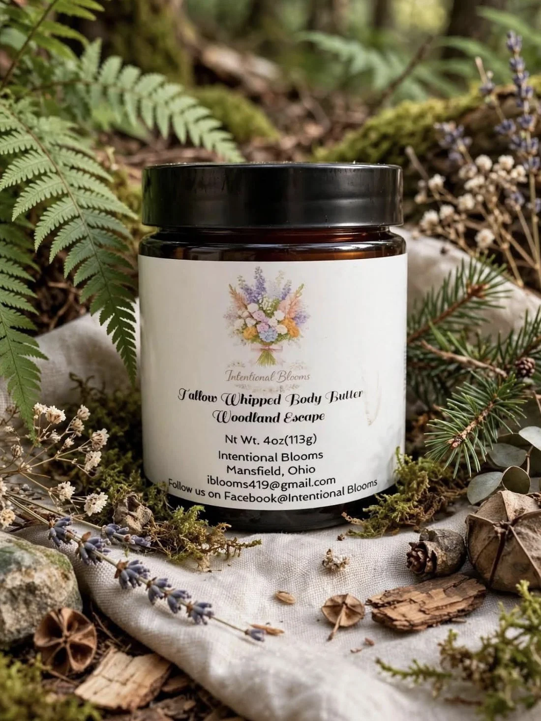 A candle jar labeled 'Tallow Whipped Body Butter Woodland Escape' from Intentional Blooms, placed outdoors on a cloth with greenery and dried flowers around it.