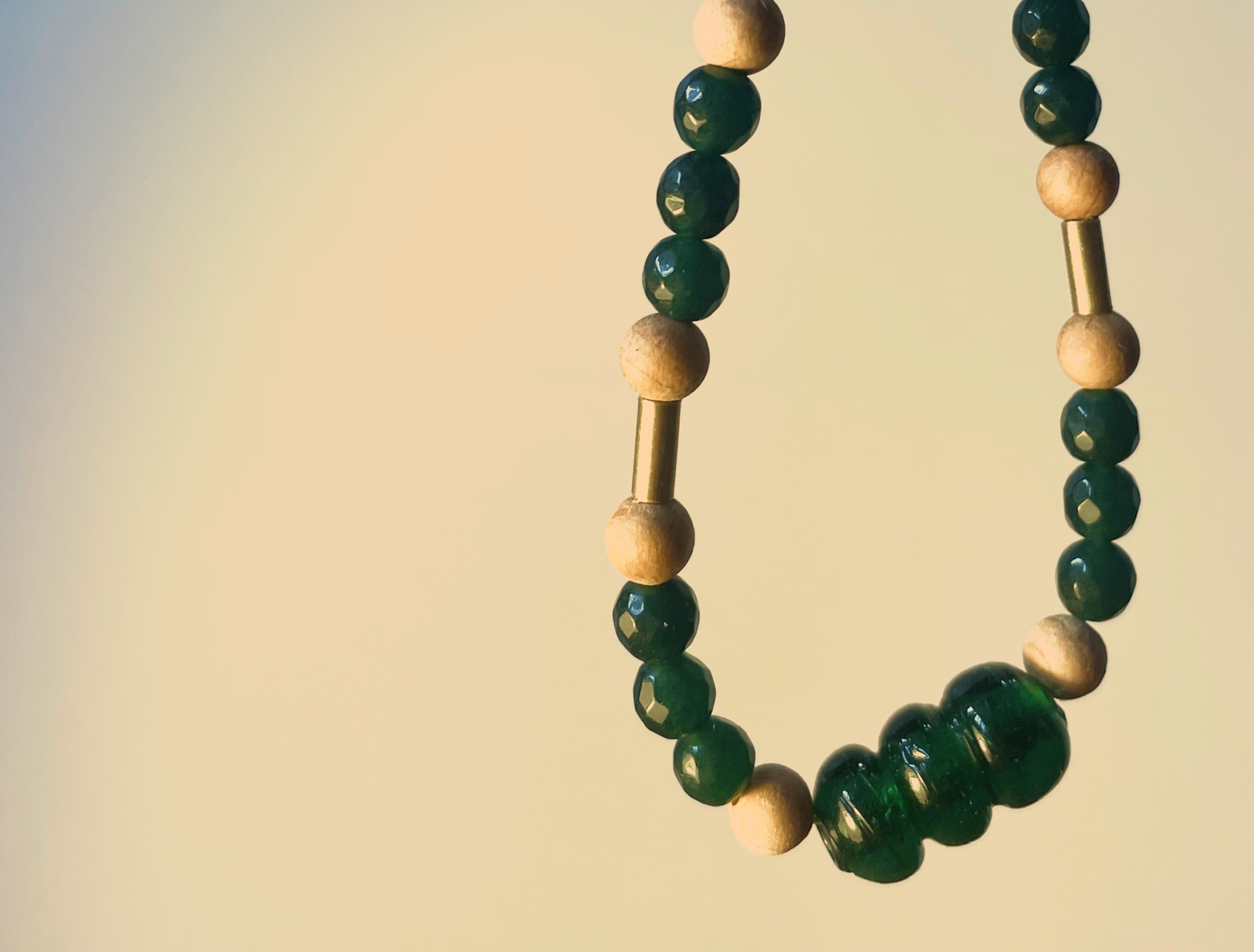 Close-up of beaded jewelry strands with dark green, cream, and metallic gold beads hanging against a neutral background.