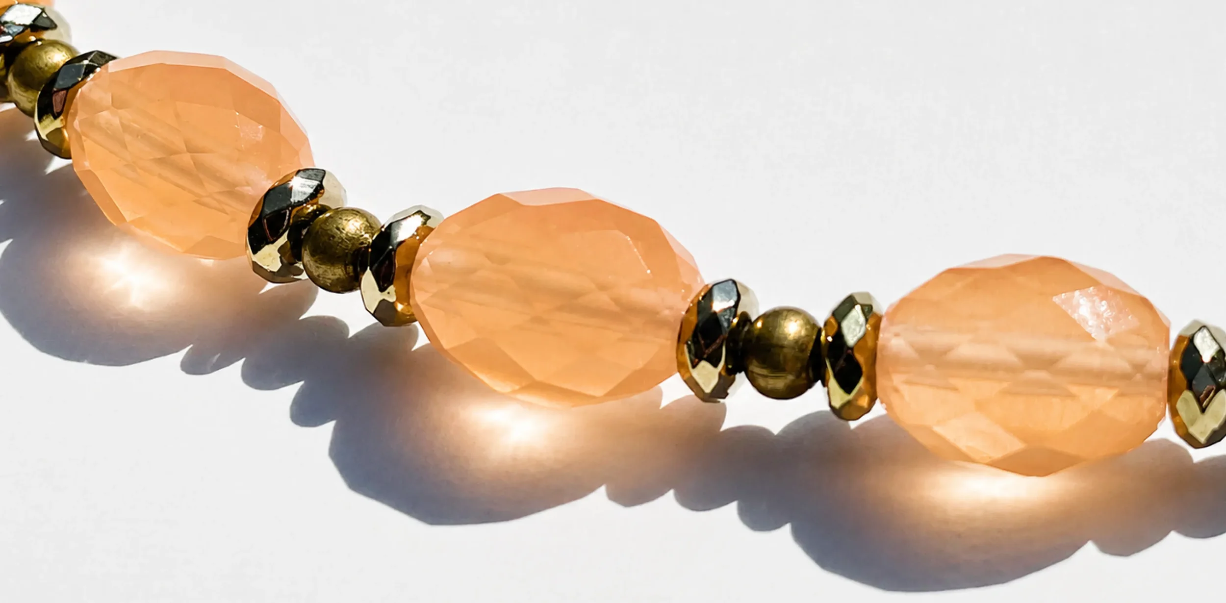 Close-up of a beaded jewelry piece with peach-colored faceted beads, metallic gold, black, and silver round beads, casting shadows on a white surface.