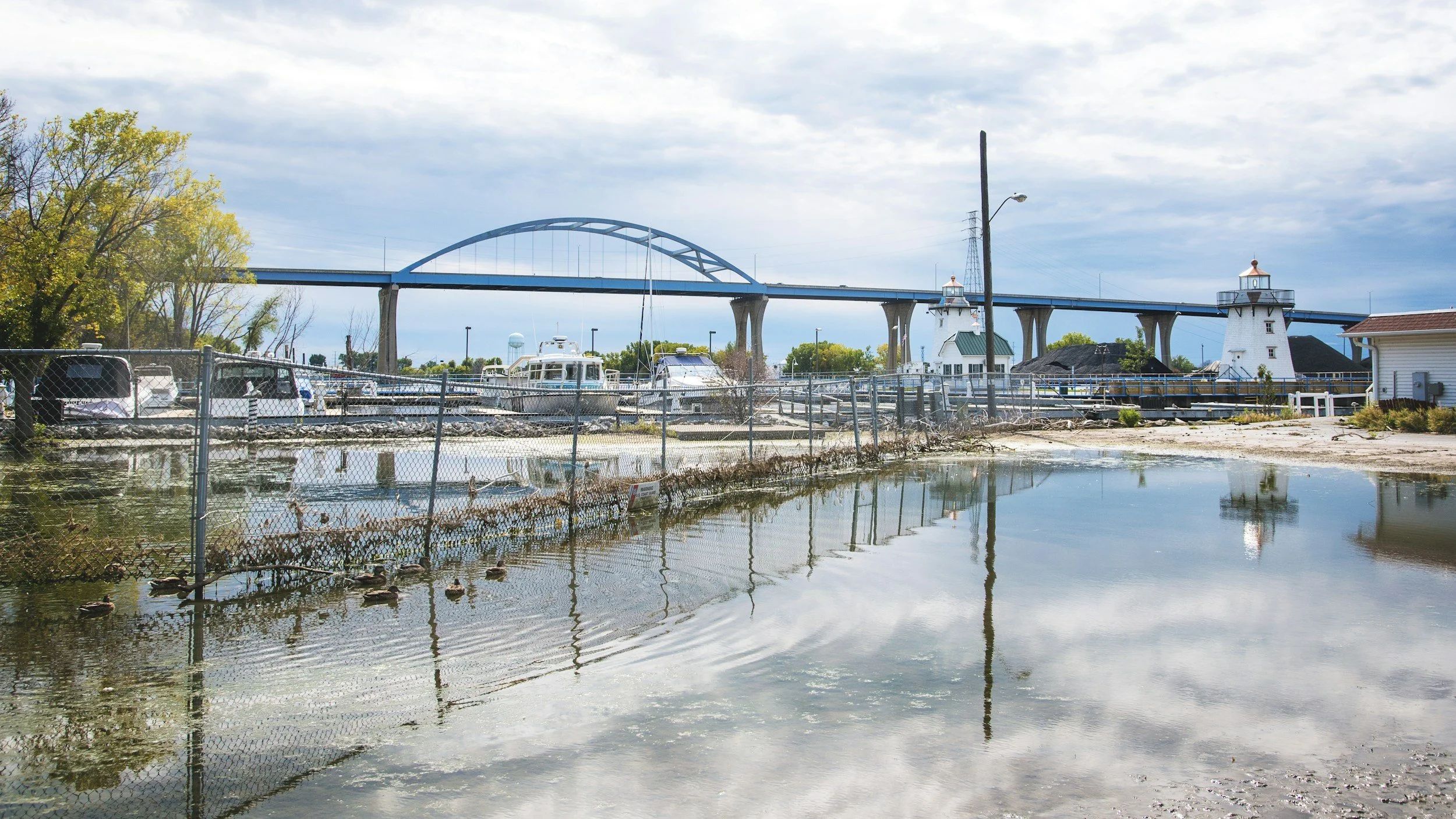 Marina with boats docked, fenced area, a large bridge in the background, cloudy sky, and reflections in the water.
