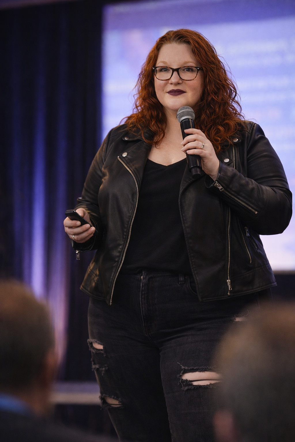 A woman with curly red hair and glasses giving a presentation on stage, holding a microphone in her right hand and a remote in her left hand, wearing a black leather jacket and ripped black jeans.