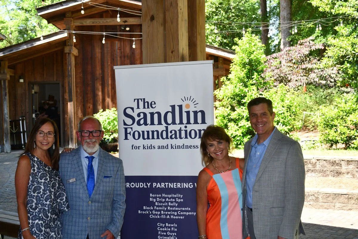 Four people standing outdoors in front of a banner for The Sandlin Foundation, smiling at the camera. The backdrop features a rustic wooden building, string lights, trees, and foliage.