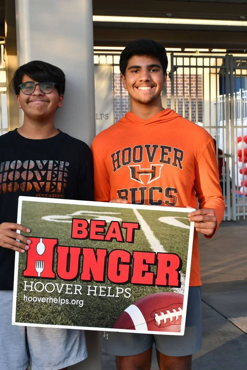 Two young men smiling at a fundraiser event. One is holding a sign that reads 'Beat Hunger' with a football and utensils graphic, promoting Hoover Helps.