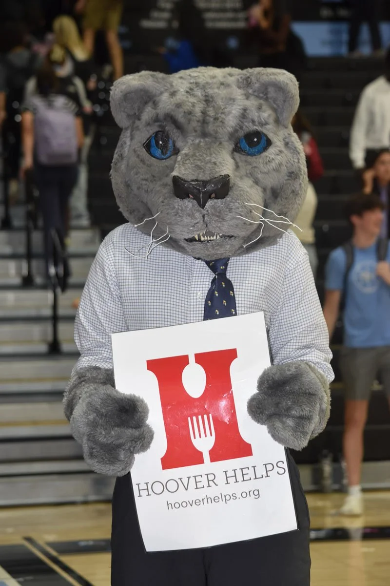 Person in a penguin or animal mascot costume wearing a business shirt and tie, holding a sign that reads 'Hover Helps' with a website link, standing in a crowded gymnasium or auditorium.