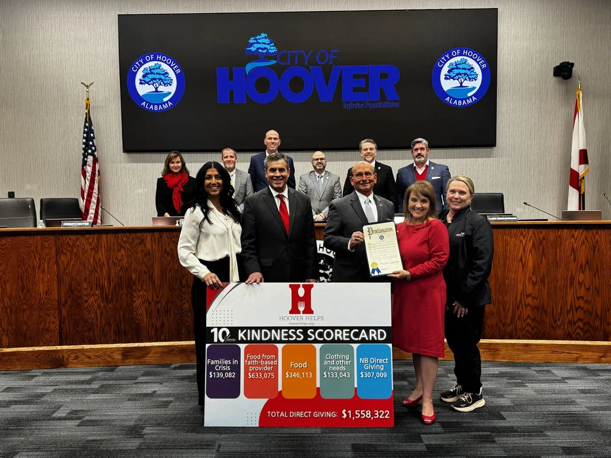 Group of people in a formal setting holding a large check and a proclamation in front of a screen displaying 'City of Hoover Alabama' and logos. The scene appears to celebrate community service or donation, with the check indicating a total of $1,558,322 in direct giving.