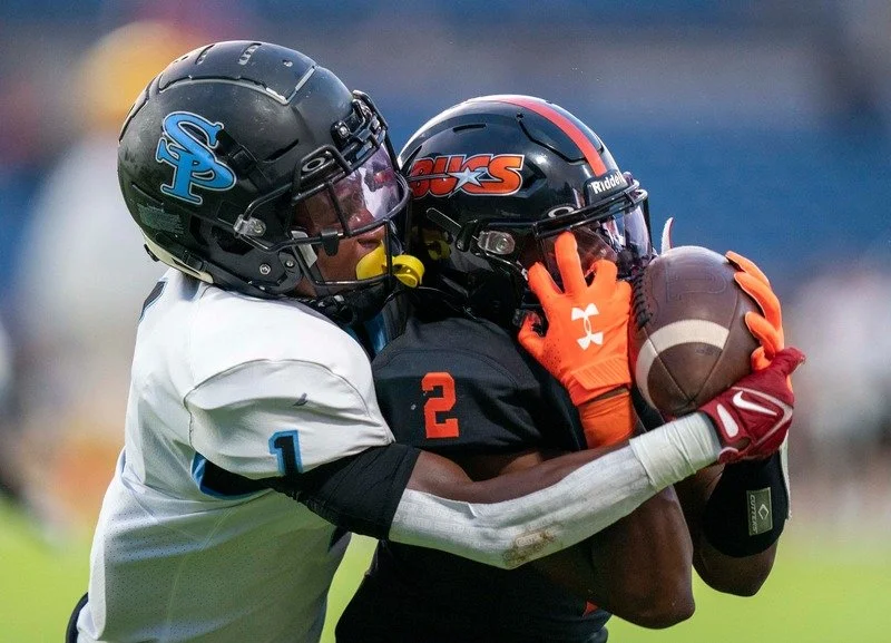 Two football players wearing helmets and gloves contest for the ball during a game.