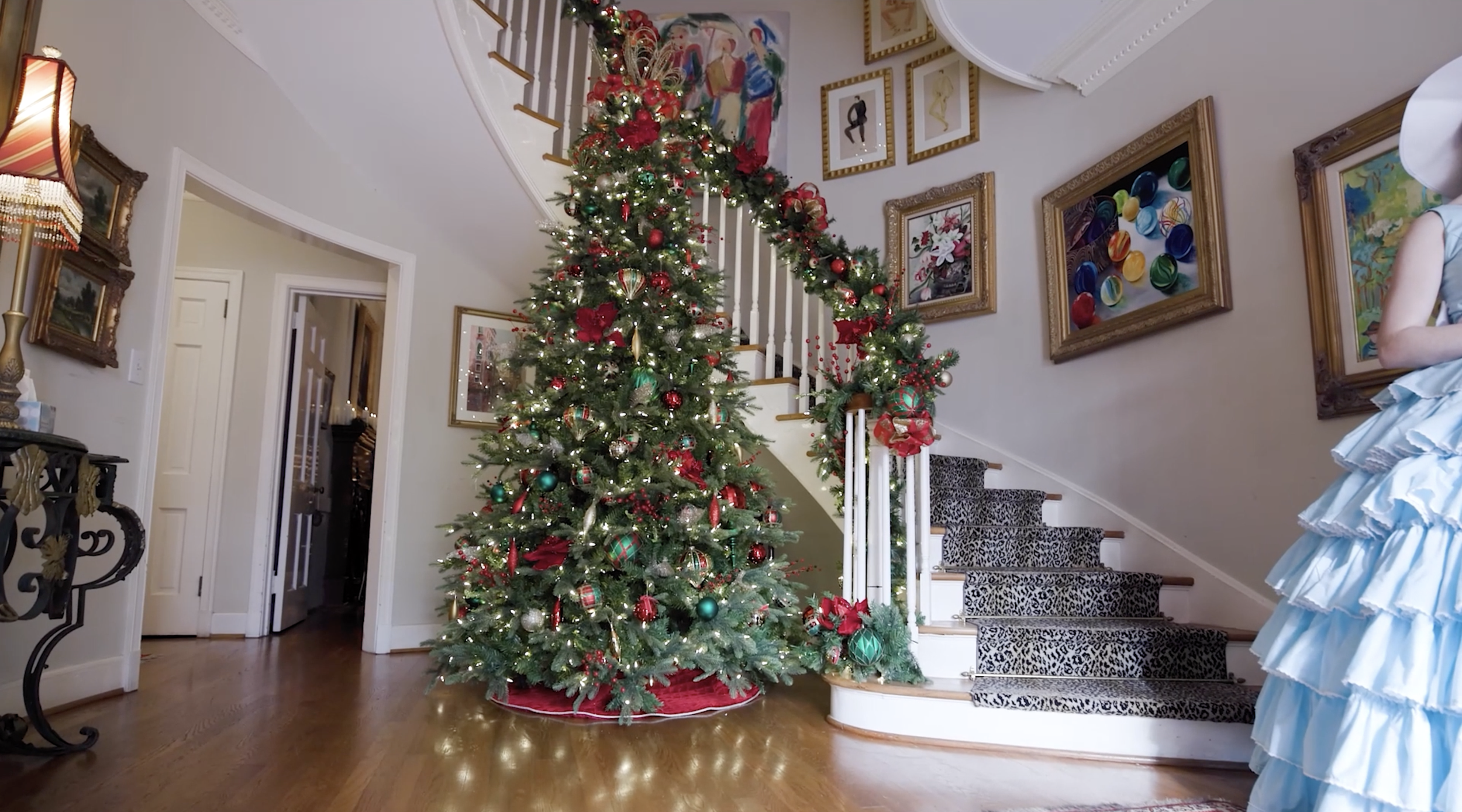 A decorated Christmas tree with red, green, gold, and silver ornaments, ribbons, and lights, placed in a living room near a staircase with garland and lights. The room has artwork on the walls, a wooden floor, and a woman in a blue dress partially visible on the right.