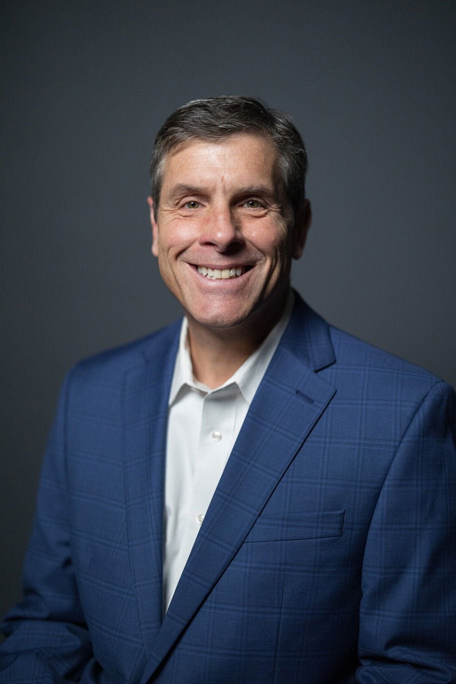 Headshot of a smiling man in a blue suit and white shirt, against a dark background.