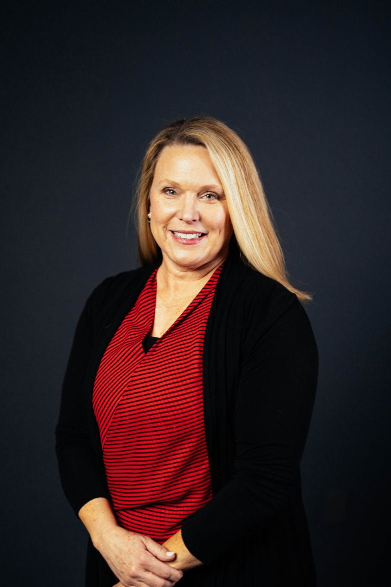 A smiling blonde woman with blue eyes wearing a red and black striped top and a black cardigan against a dark background.