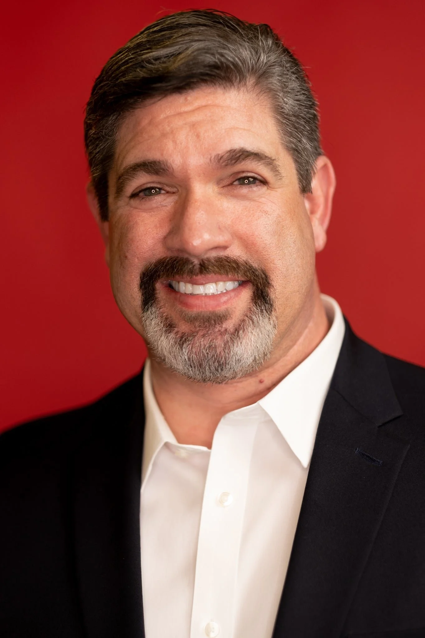 A man with gray hair and a beard, wearing a black tuxedo jacket and white dress shirt, smiling against a red background.