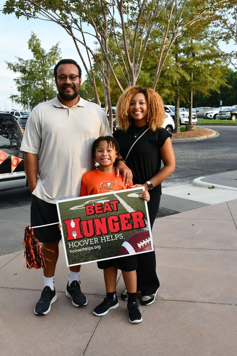 A smiling family of three standing outdoors in a parking lot, holding a sign that reads 'Beat Hunger, Hoover Helps' with a football graphic, promoting a hunger relief campaign.