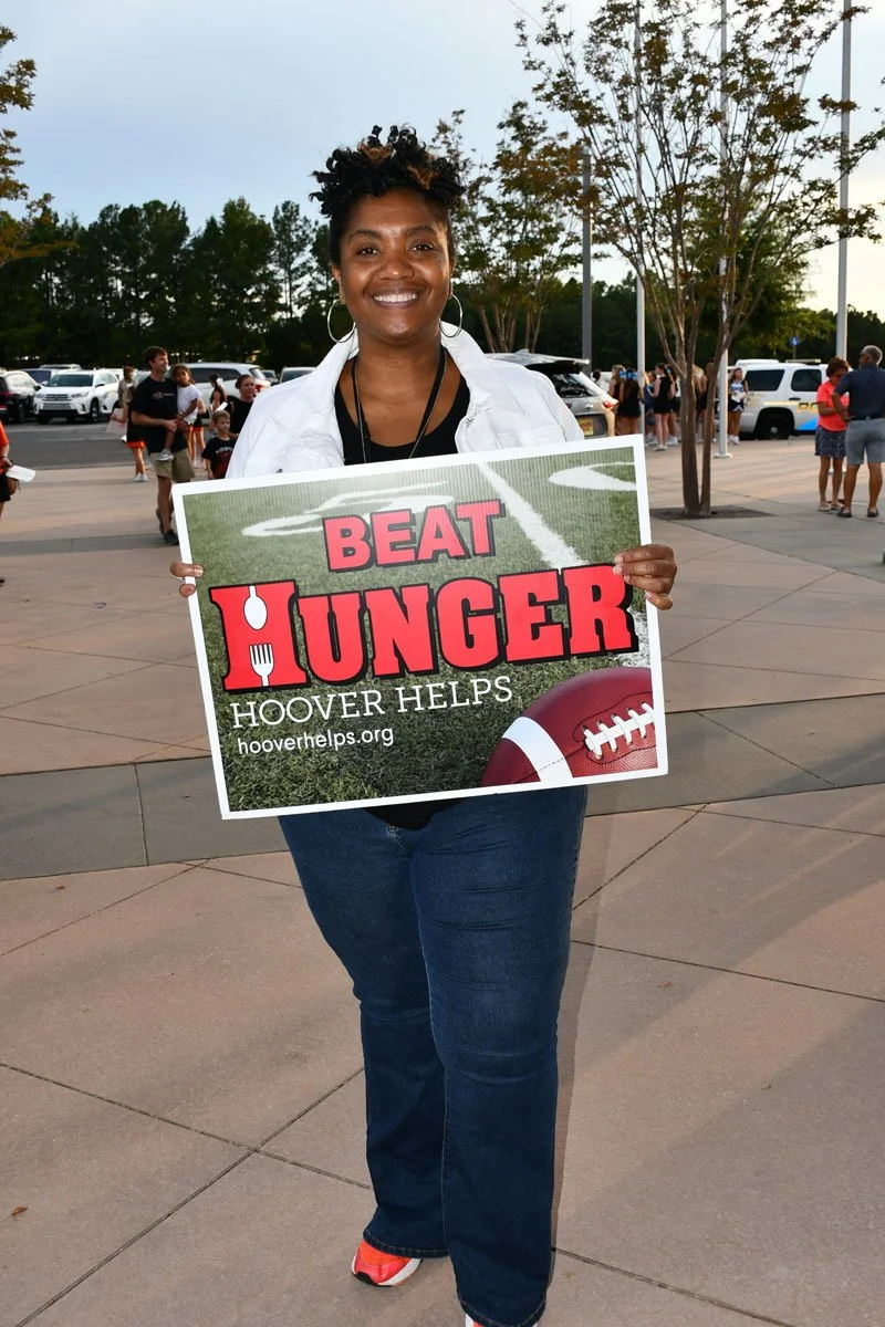 Smiling woman holding a sign promoting Hoover Helps to beat hunger, with a football graphic, outdoors with trees and parked cars in the background.