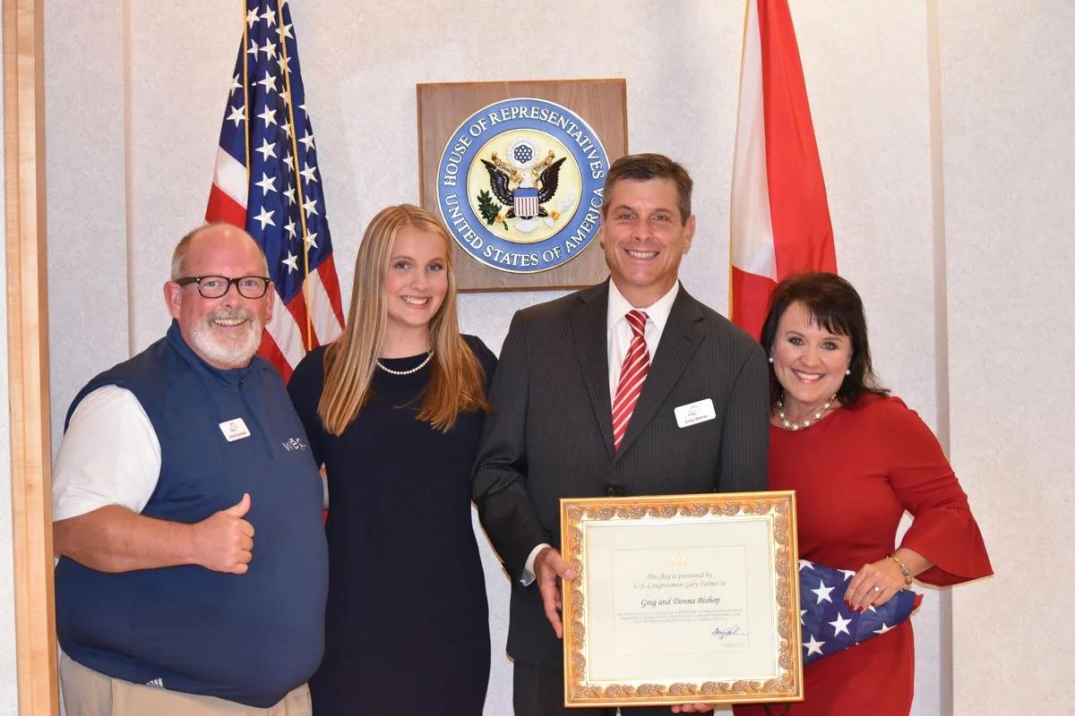 Four people standing together in front of a U.S. House of Representatives seal, with American and Canadian flags behind them. One man in a suit holds a framed certificate, and they are all smiling.
