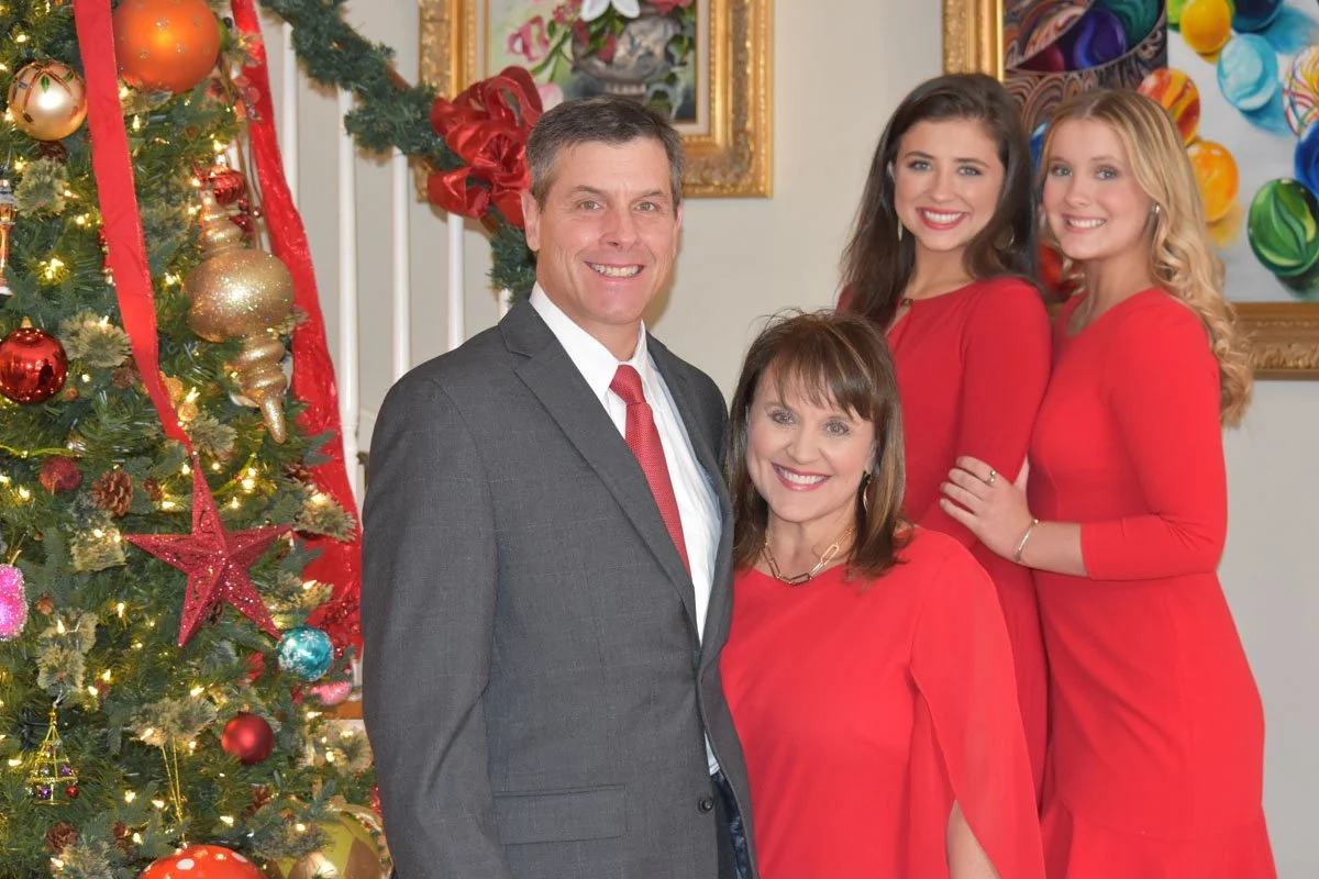 A family dressed in red posing in front of a decorated Christmas tree with ornaments, lights, and ribbons in a decorated room.