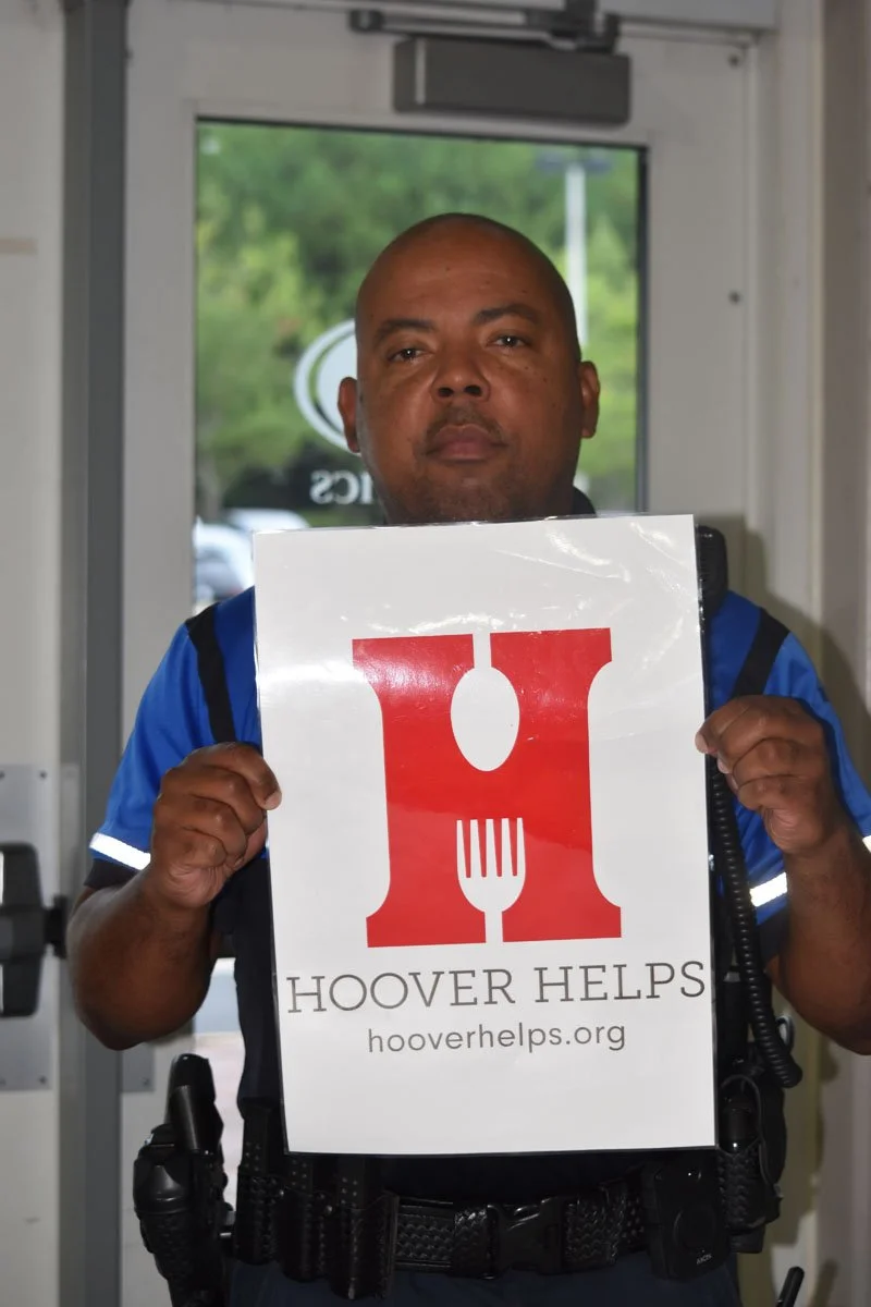 A man in a blue uniform holding a sign with a red logo and text that reads 'HOOVER HELPS' and the website hooverhelps.org, standing inside near a door with a view of greenery outside.