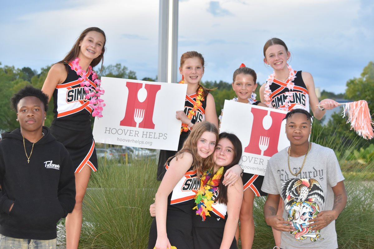 Group of eight young girls and boys outdoors holding cheer signs that say "Hoover Helps" with a logo of a fork, spoon, and plate. The girls wear cheerleading uniforms with "Simpson" on them, and some have leis.