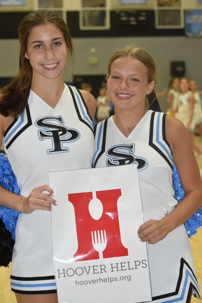 Two young women in cheerleading uniforms holding a sign for Hoover Helps organization in a gymnasium.