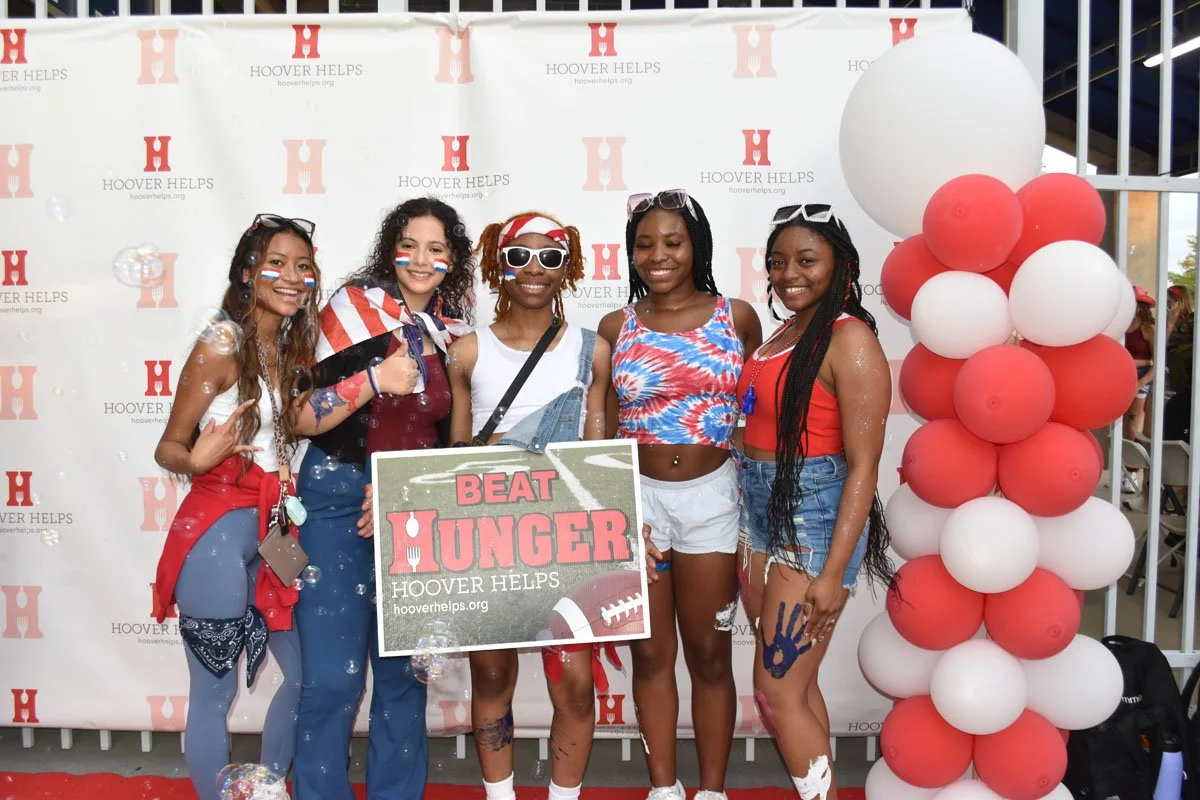 Group of six young women dressed in casual summer clothes, celebrating at an event, holding a sign that says 'Beat Hunger' with balloons and a branded step-and-repeat backdrop in the background.