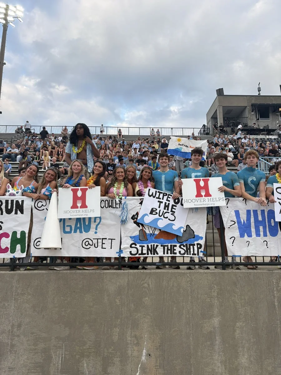 A group of teenagers standing behind a fence at a stadium, holding colorful signs and banners with humorous messages and logos, some wearing leis and summer attire.
