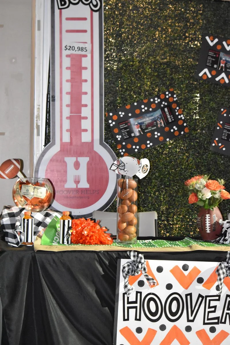 A football-themed party table with a large thermometer sign indicating a donation amount of $20,985, decorations including footballs, orange and black pom-poms, and framed flyers with orange, black, and white patterns. There are flowers in a football