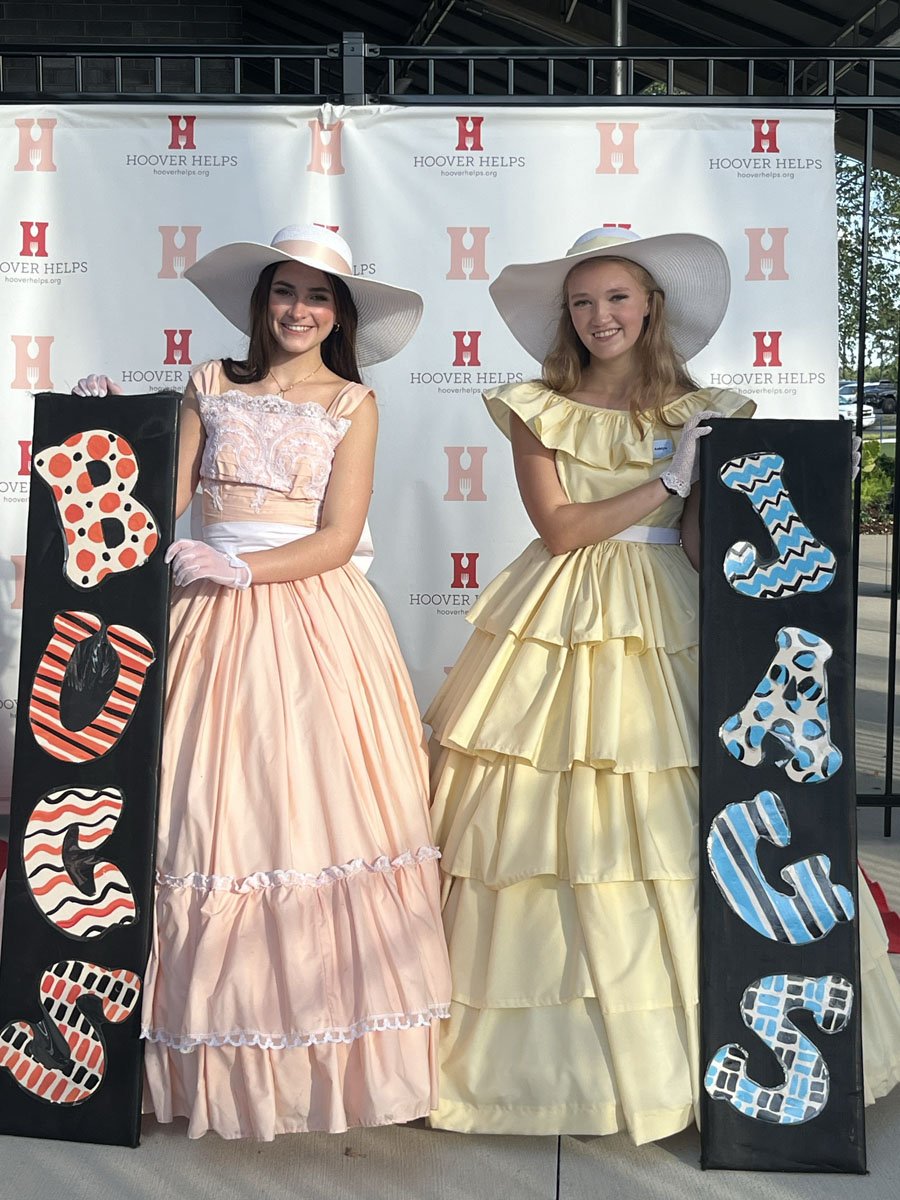 Two young women dressed in vintage-style dresses and large sun hats holding black boards with colorful, patterned letters that spell "BOYS" and "GIRLS". They are standing in front of a step-and-repeat backdrop with the Hoover Helps logo.