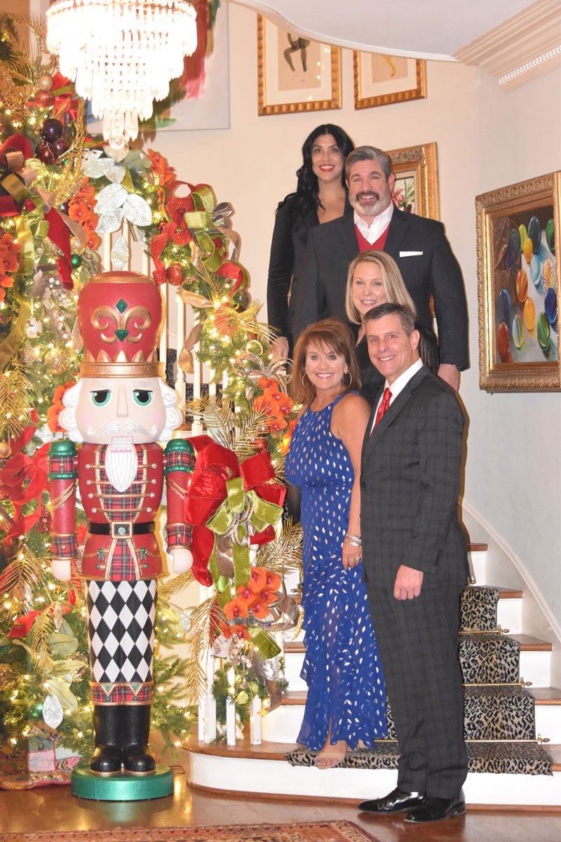 Group of five people, three women and two men, standing on a staircase decorated for Christmas with a large Christmas tree and a nutcracker figure nearby.
