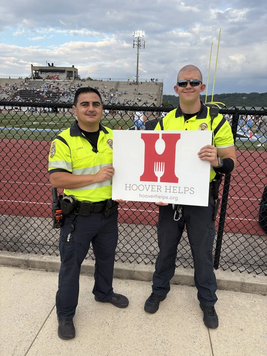 Two police officers standing in front of a football field fence, holding a sign that says 'HOOVER HELPS' with a logo of a red plate, fork, and knife, at an outdoor event.