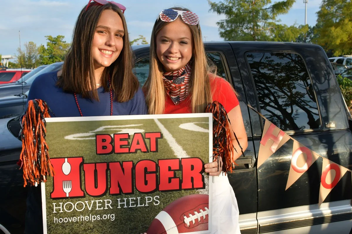 Two young women smiling and holding a sign that says "Beat Hunger" and "Hoover Helps," standing in front of a parked vehicle with a patriotic banner and a football on it.