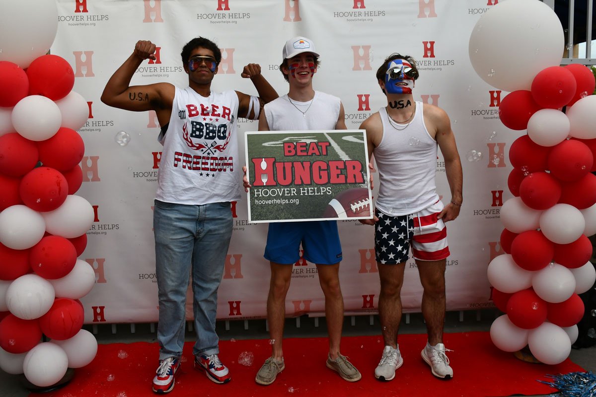 Three young men in patriotic clothing and face paint, posing with fists raised and a sign that reads 'Beat Hunger' at an event with red and white balloon decorations and a backdrop with the Hoover Helps logo.