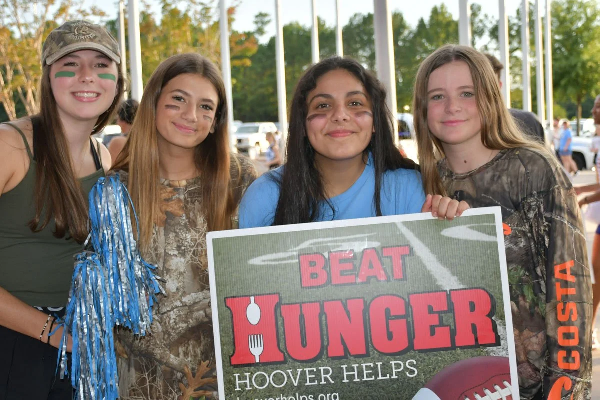 Four young women smiling, standing outdoors, holding a sign that says "BEAT HUNGER, HOOVER HELPS." They are dressed in casual and camouflage clothing, with some wearing face paint and one holding blue and silver streamers.