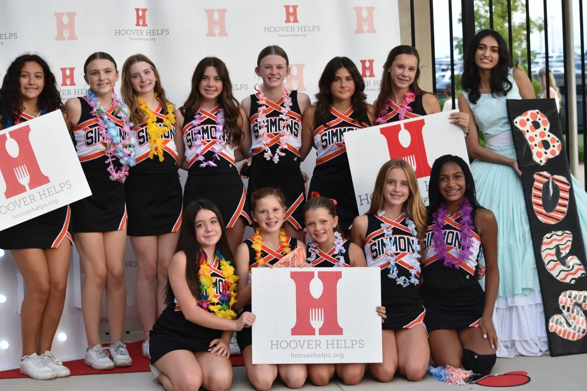 Group of young girls in cheerleading uniforms holding signs for Hoover Helps, with a woman dressed as a cake on the side.