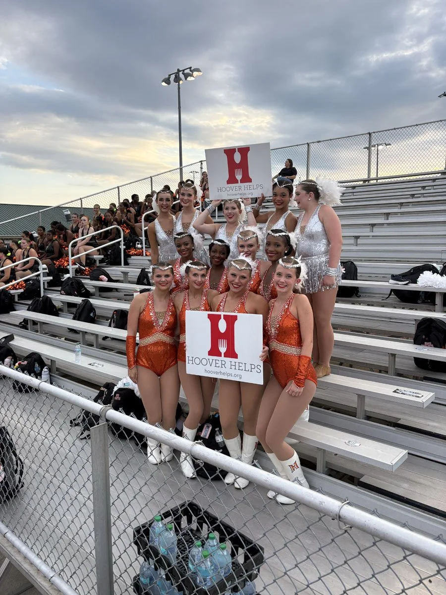 A group of cheerleaders in orange and white uniforms holding signs that say "HOOVER HELPS" at an outdoor sports stadium.