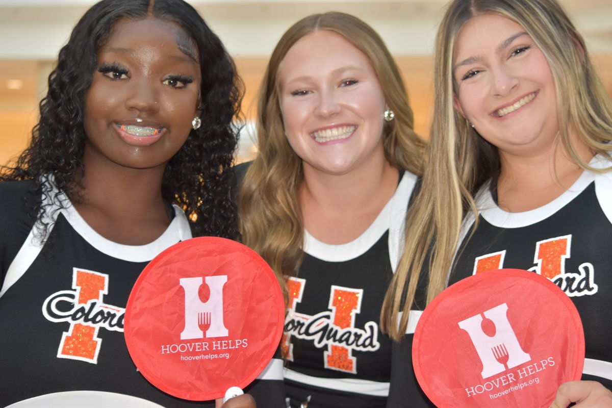 Three young women wearing matching black and white sports jerseys with orange 'H' and 'Color Guard' text, smiling, at an indoor event, holding red handheld fans with white 'H' and 'Hoover Helps' logo.
