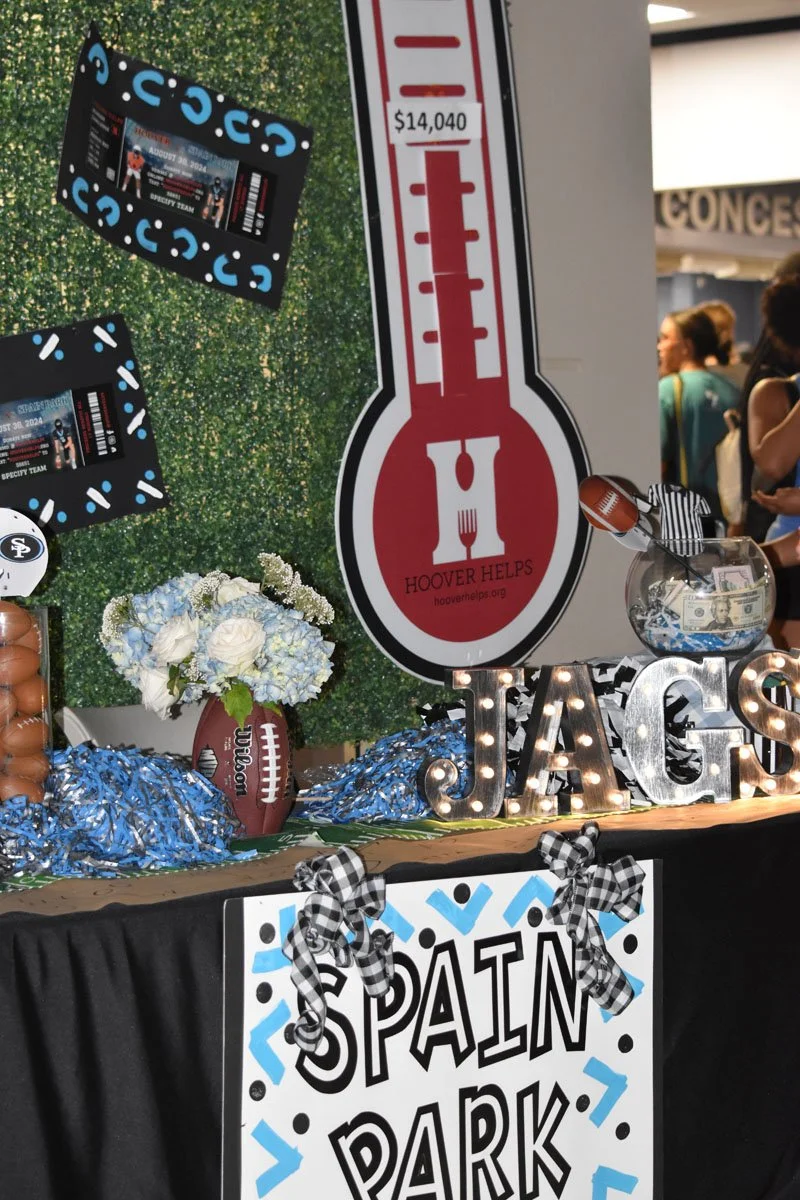 A decorated table at a sports-themed event featuring a sign that says "Spain Park" with black and white bow decorations, a large thermometer graphic indicating $14,040, a glass jar with cash and a football, and a bouquet of white and blue flowers, al