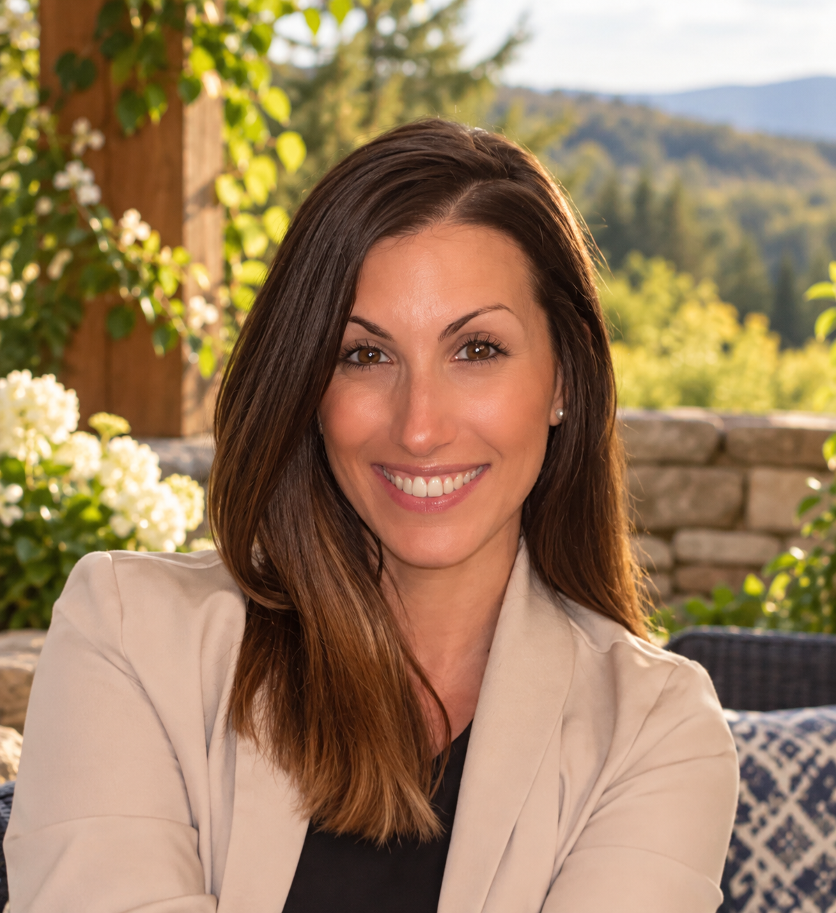 A smiling woman with brown hair and brown eyes, wearing a beige blazer and a black top, sitting outdoors with a background of trees, flowers, and mountains.