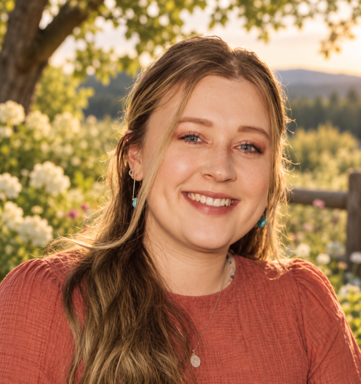 Smiling woman with long wavy hair wearing earrings and a necklace outdoors during sunset, with trees and mountains in the background.