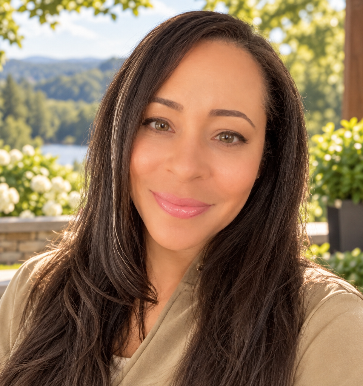 Close-up of a woman with long dark brown hair, smiling, outdoors with trees, flowers, and a lake in the background.