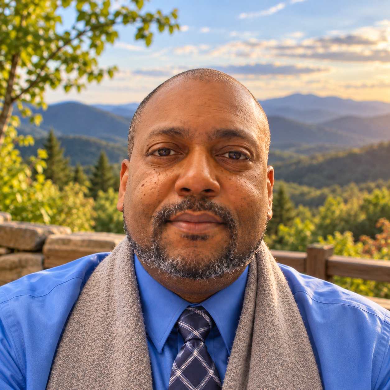 Close-up of a middle-aged man with a gray beard, wearing a blue dress shirt, a patterned tie, and a gray scarf, outdoors with a scenic mountain landscape and a partly cloudy sky in the background.