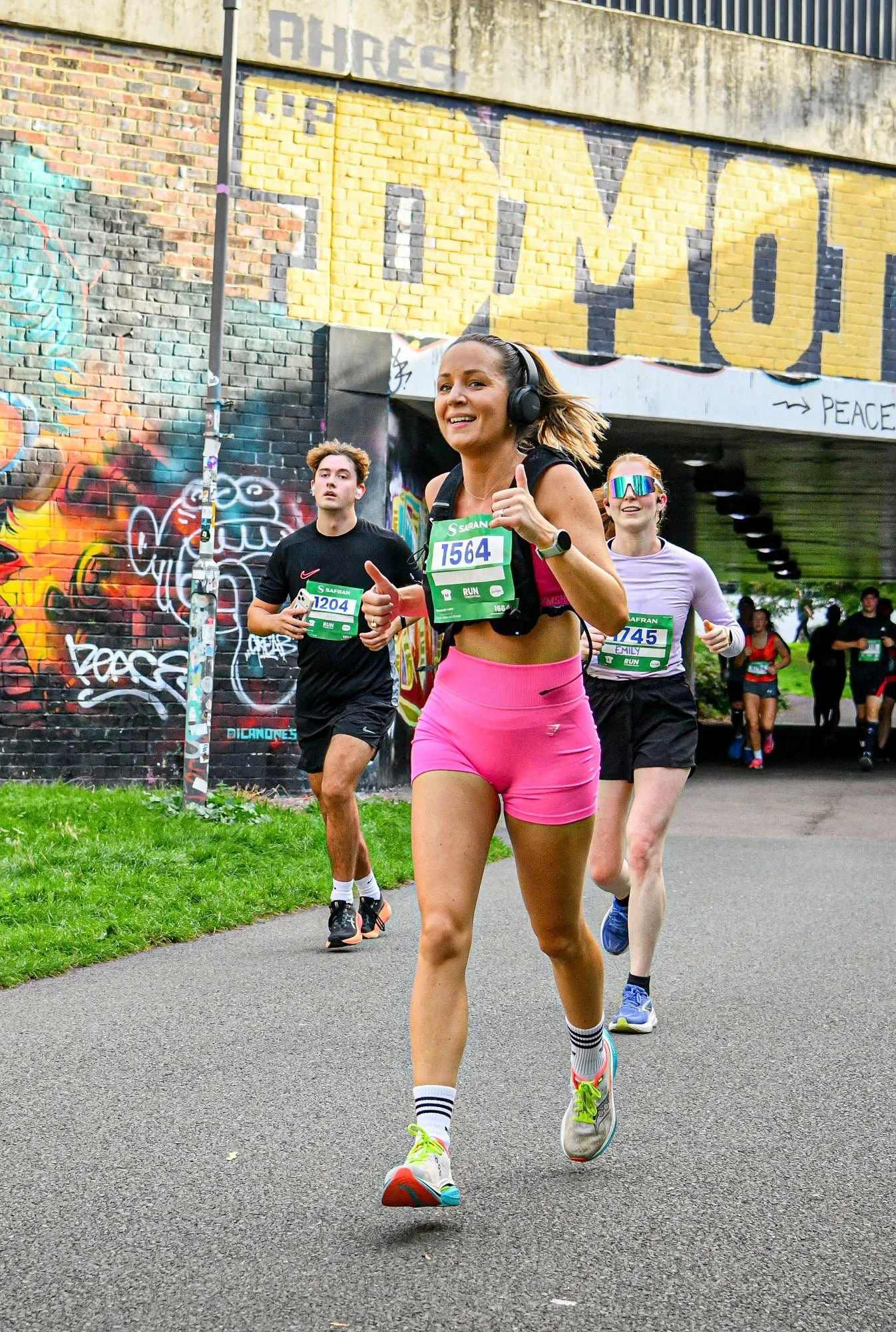 A group of runners participating in a marathon or race, running along a paved path under a graffiti-covered bridge. The lead runner, a woman wearing pink shorts, a black sports bra, and neon yellow running shoes, is smiling and wearing headphones. Other runners are visible behind her, some with race bibs and athletic clothing.
