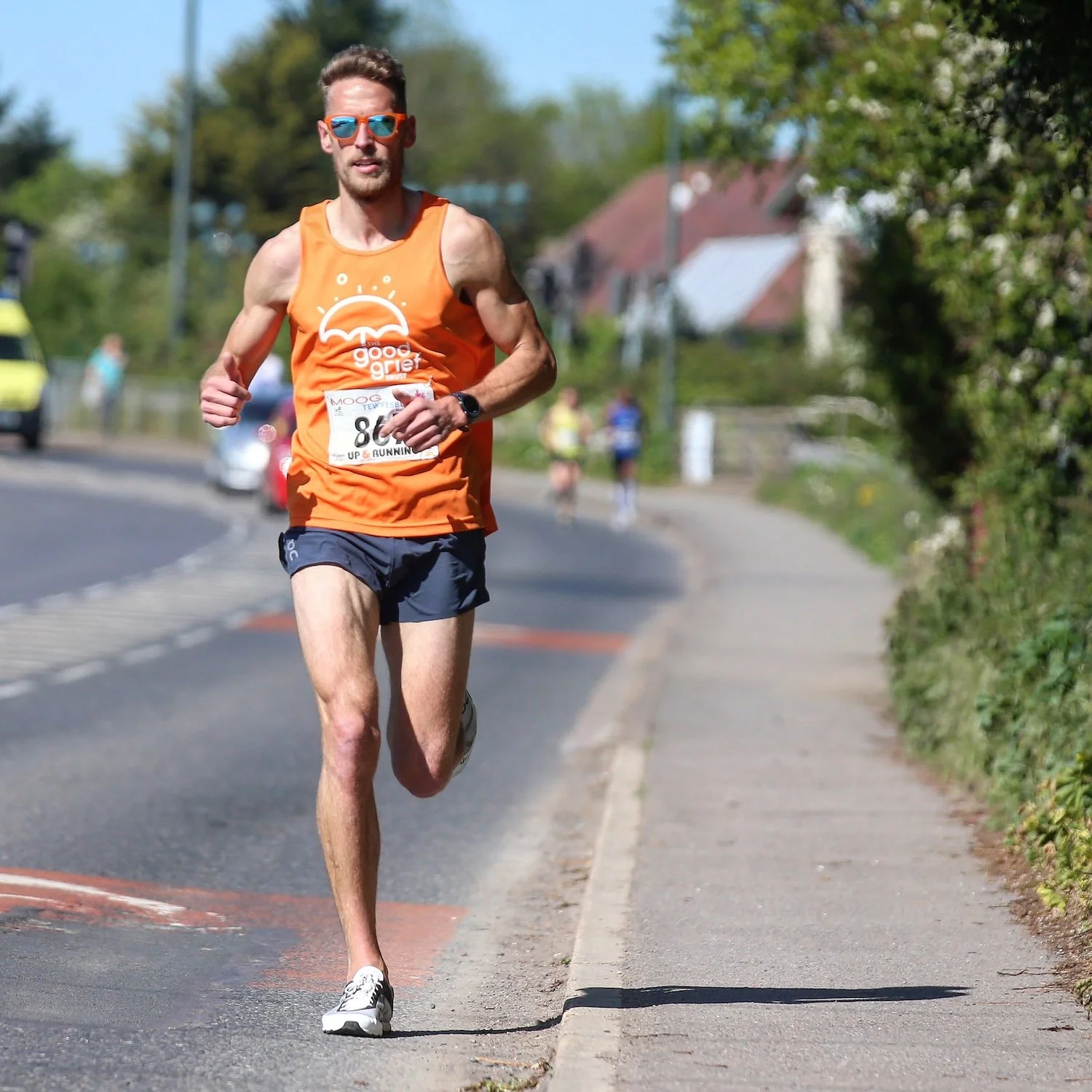 Man running in a marathon wearing sunglasses, an orange sleeveless shirt with a logo, and black shorts, on a sunny day.