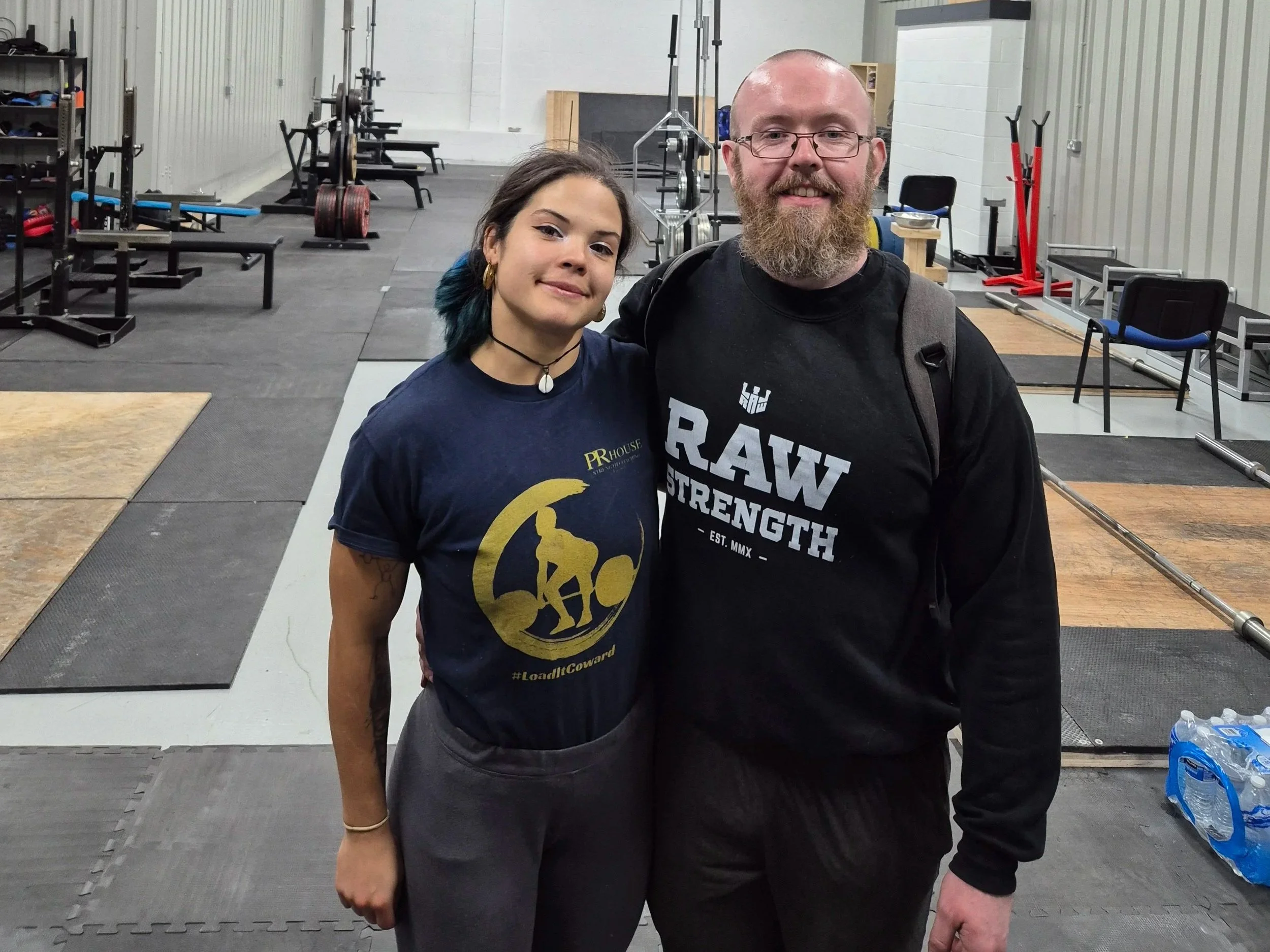 Two people standing together in a gym or fitness facility, smiling at the camera. The woman has dark hair with blue tips, wearing a navy T-shirt with a gold logo and necklace. The man has a full beard, glasses, and is wearing a black T-shirt with white text and a backpack. The background shows exercise equipment, weights, benches, and a black rubber floor.