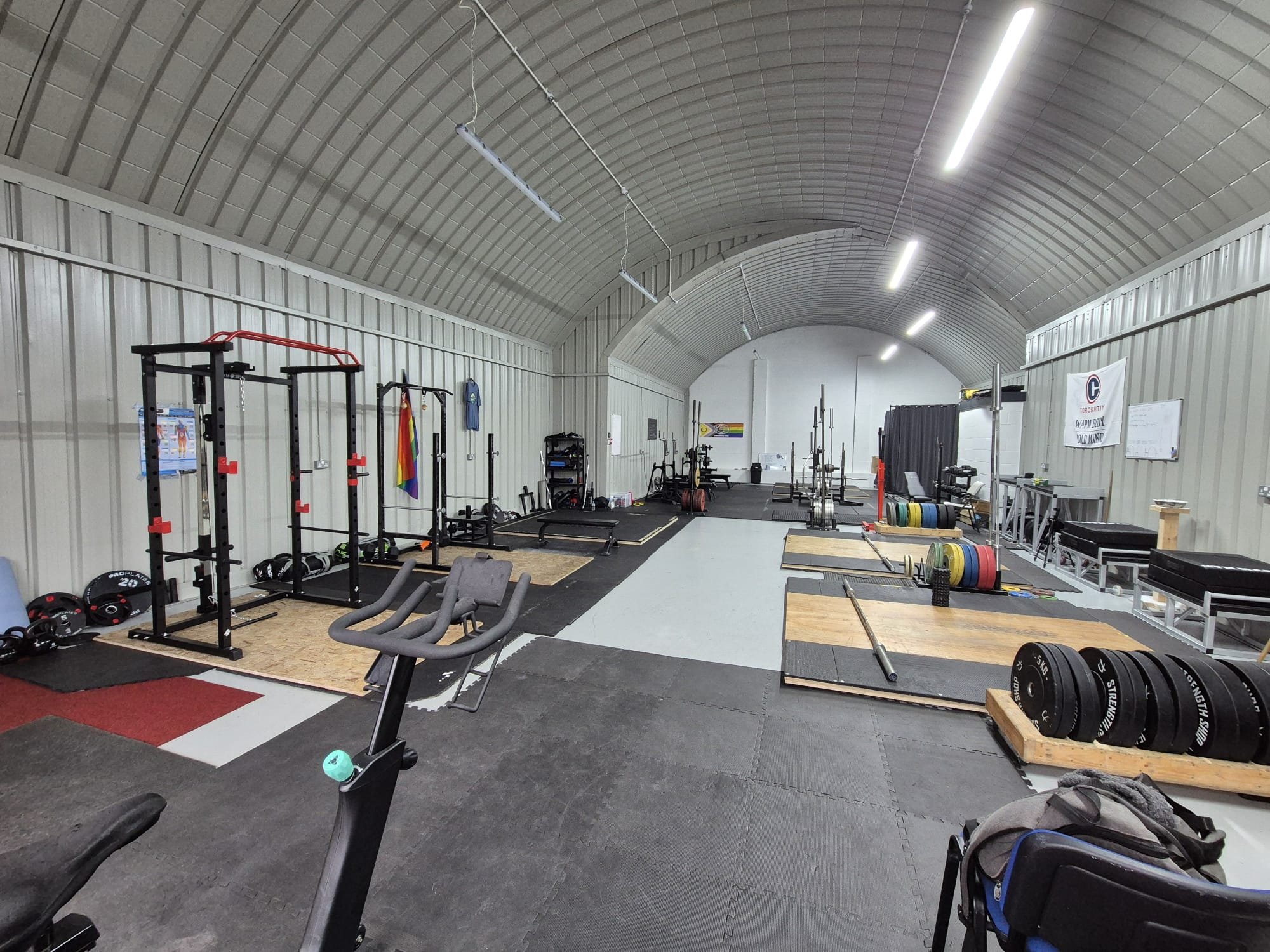 Inside a spacious gym with high, curved metal ceiling and bright overhead lighting. The gym has various workout equipment including squat racks, weight plates, barbells, and benches. A rainbow pride flag and gym-related signs are visible on the walls.