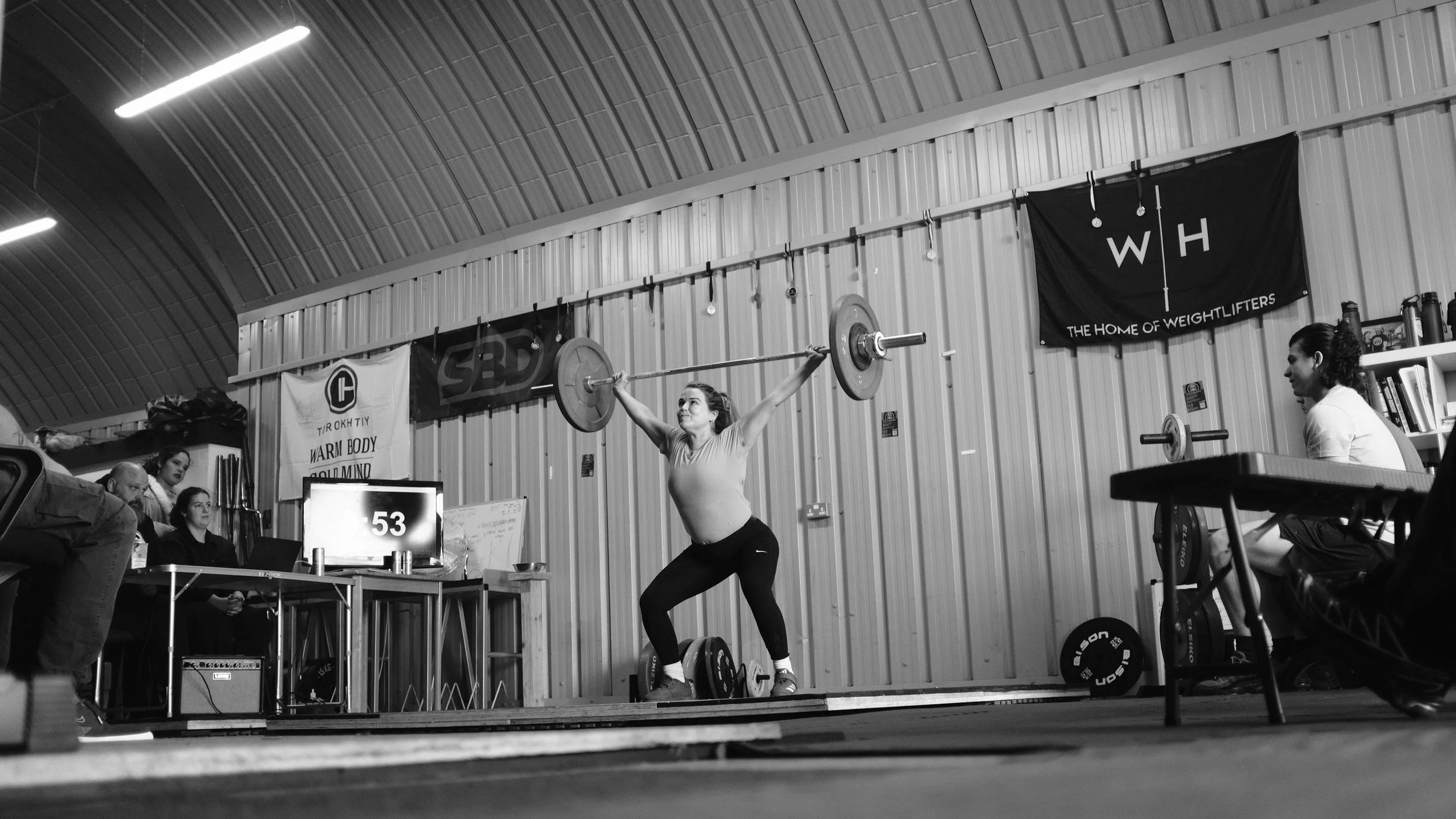 A woman weightlifter is performing a clean and jerk at a weightlifting competition, with her arms extended overhead holding a barbell with weights. Several spectators and officials are watching around her in a gymnasium with banners on the wall.