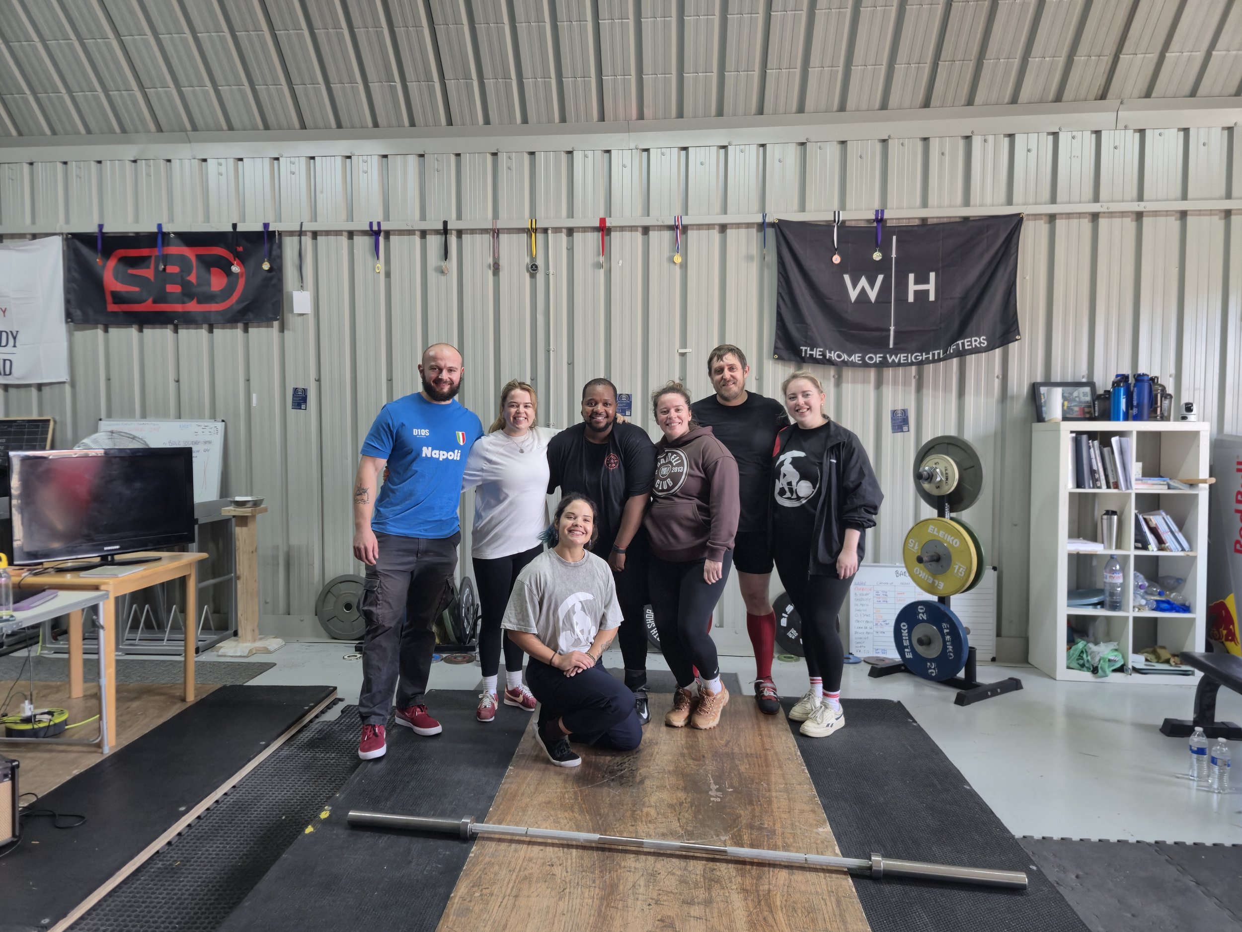 Group of six people standing and kneeling inside a gym with weightlifting and fitness equipment, including weight plates, a whiteboard, banners, and water bottles.