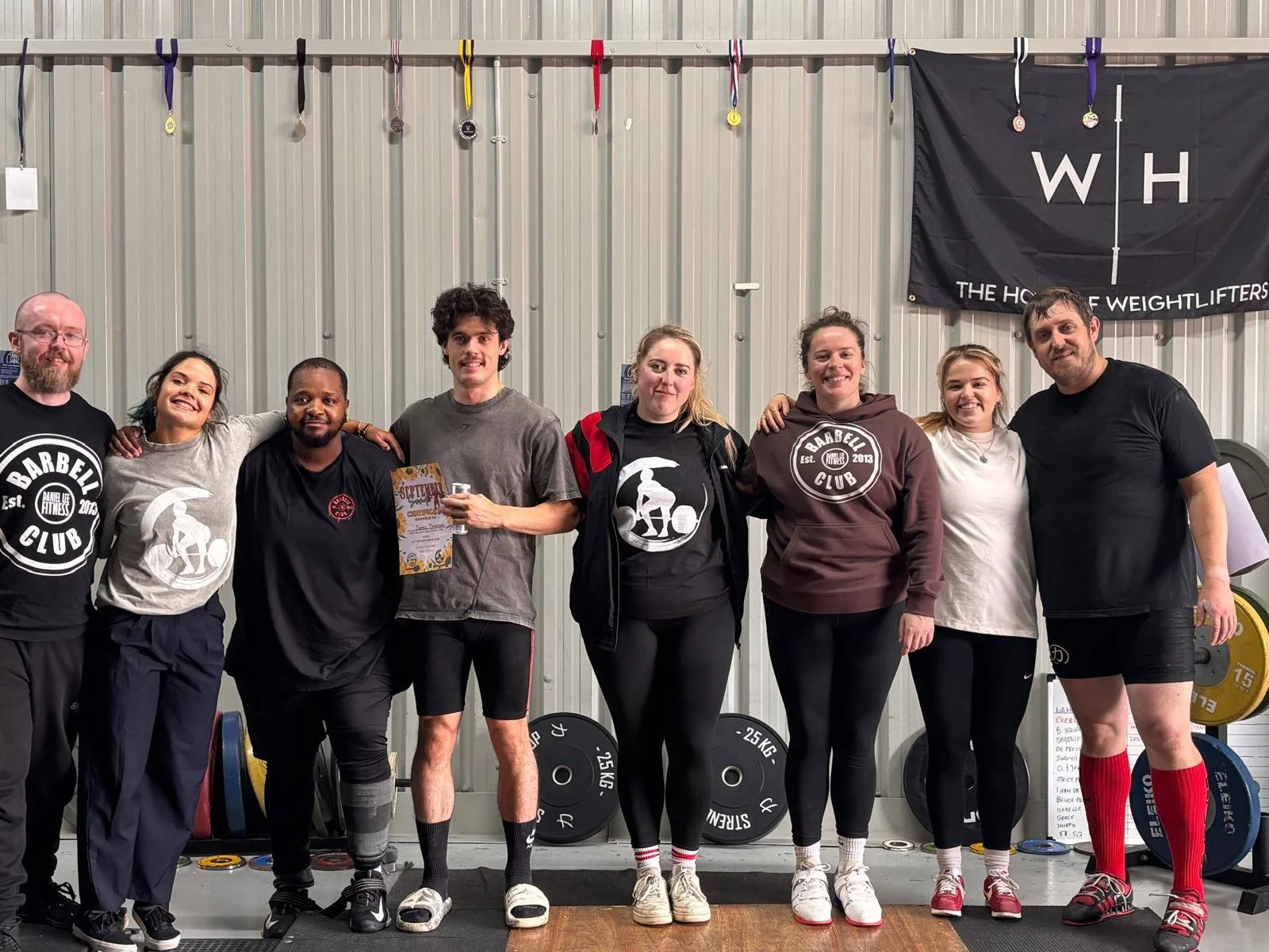 Group of eight people standing together in a gym, some holding medals, with weights behind them and a banner that reads "The House of Weightlifters".