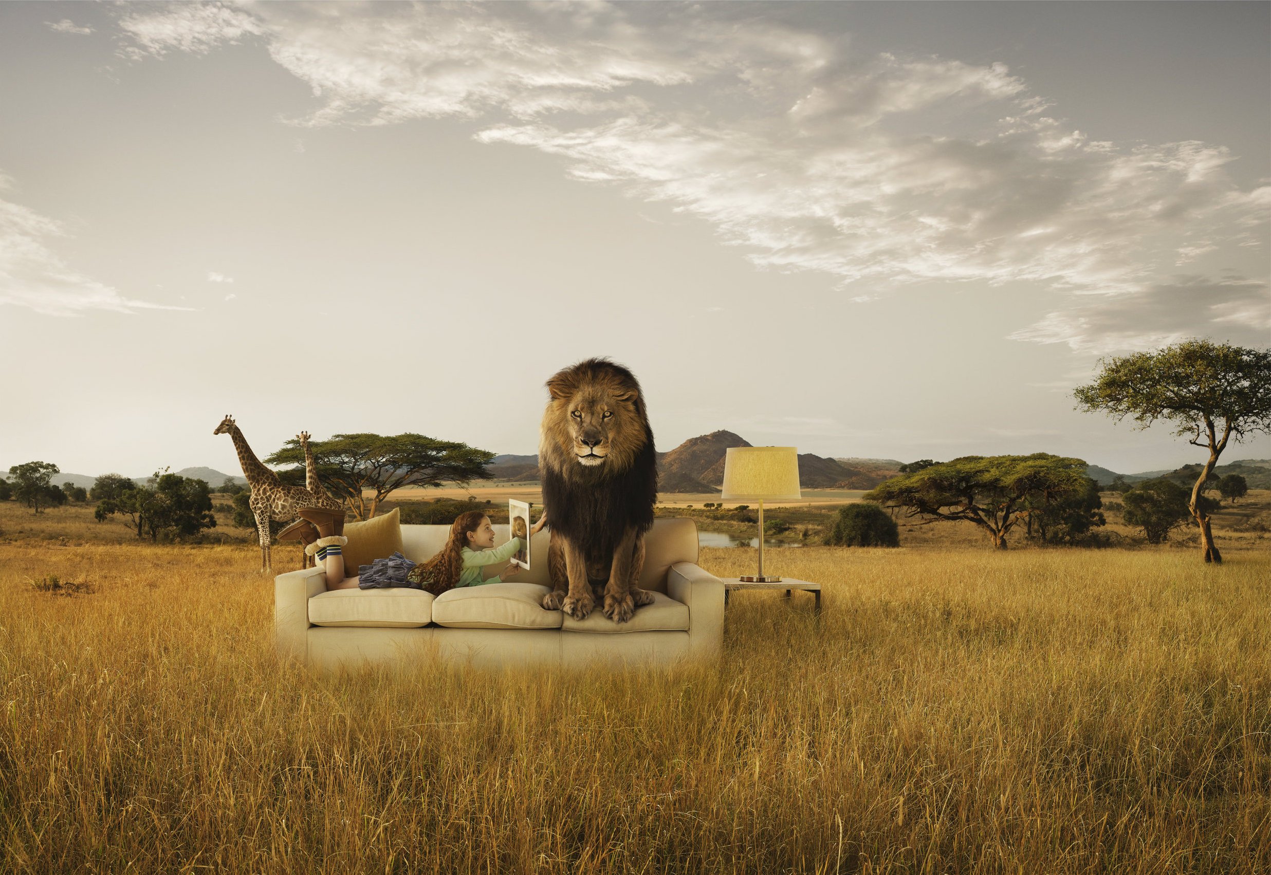 A girl with red hair sits on a beige sofa outdoors in a savannah landscape with giraffes, a lion statue, and trees under a cloudy sky.