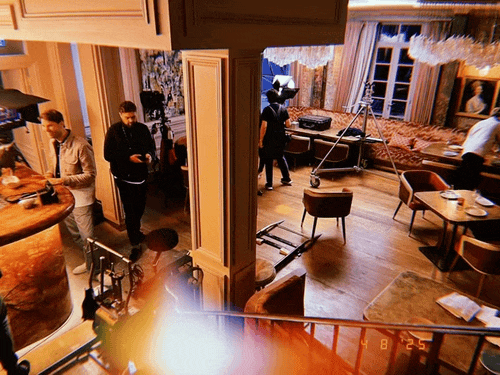 People in a spacious, well-decorated room with wooden floors, dining tables, and chairs, possibly a restaurant or café, with some individuals standing and chatting, and others walking around.
